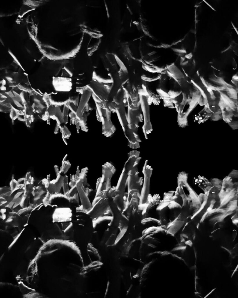 Black and white, mirrored  image of raised hands in a concert. Among them a mobile is catching the scene