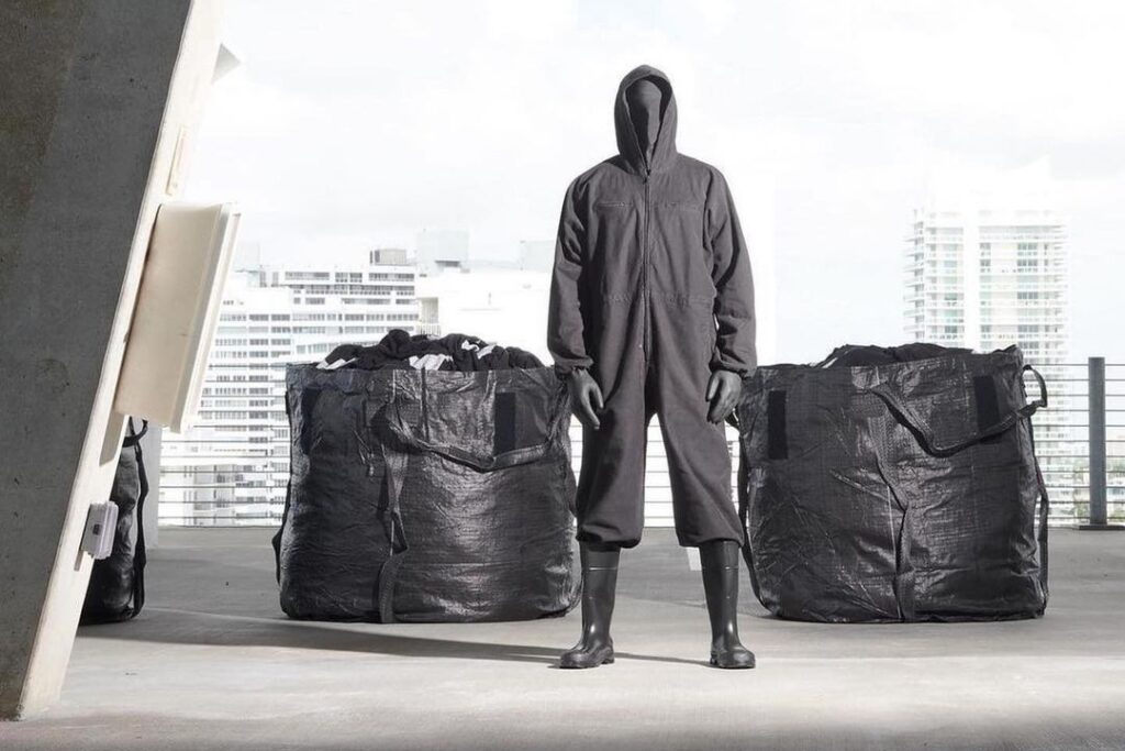 A model wearing Yeezy x Gap collection is standing between two big black trash bags